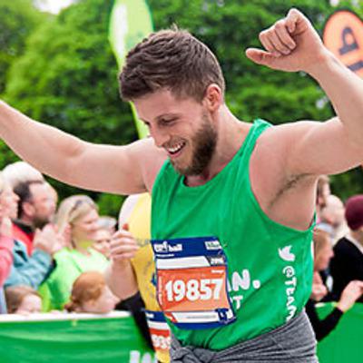 A runner, wearing a green tank top and bib number 19857, rejoices with both arms raised at the finish line of a race. A crowd of spectators claps and cheers in the background. Bright greenery is visible behind the crowd.