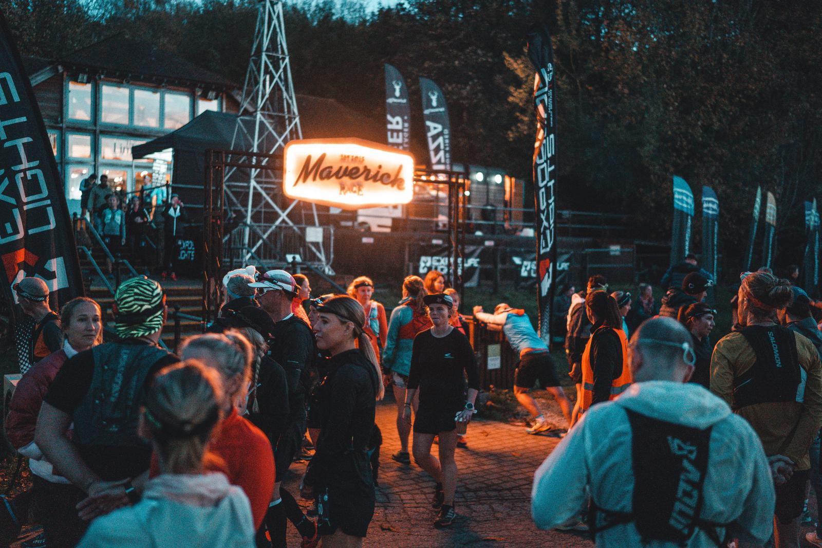 A diverse group of people gather in front of a building at dusk, illuminated by a bright neon sign reading "Maverick." Many are wearing athletic gear and appear to be participating in an outdoor event or race, with banners and flags visible in the background.