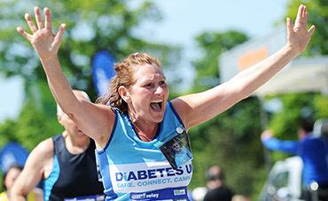 A joyful runner in a blue "Diabetes UK" tank top, with outstretched arms, celebrates while finishing a race in outdoor summer conditions. The runner appears to be elated with their accomplishment. Other runners and greenery are seen in the background.