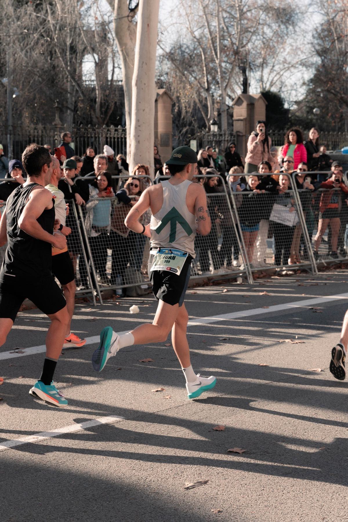 A group of runners competing in a street race, with spectators behind a metal fence on the side of the road. One runner in the foreground wears a gray tank top with a green arrow design, black shorts, and a green cap. Trees and buildings are visible in the background.