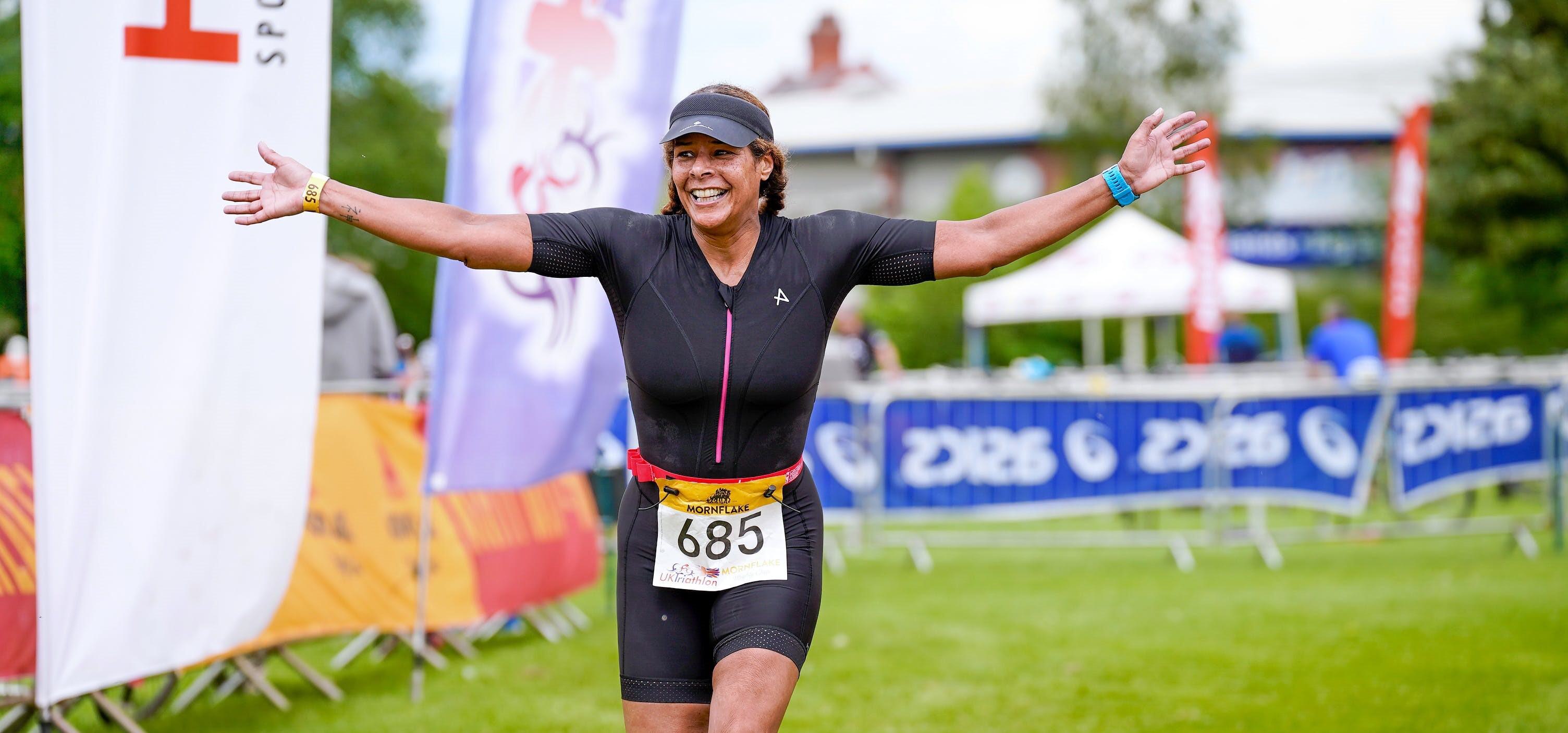 A person wearing a black triathlon suit and race bib number 685 smiles broadly with arms outstretched while crossing the finish line at an outdoor event. In the background, there are banners, spectators, and a grassy area.