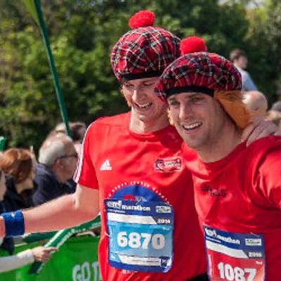 Two men in red T-shirts and checkered hats with red pom-poms, smiling and posing for a photo at a marathon event. One holds a smartphone, and both have race numbers pinned to their shirts. People and trees are visible in the background on a sunny day.