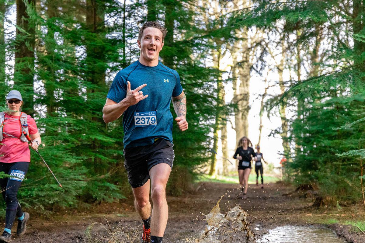 A man wearing a blue shirt and black shorts runs and smiles, making a hand gesture, through a forest trail. His race bib number is 2379. Other runners can be seen in the background, with one wearing a pink shirt and hat. The ground is muddy with water splashes.