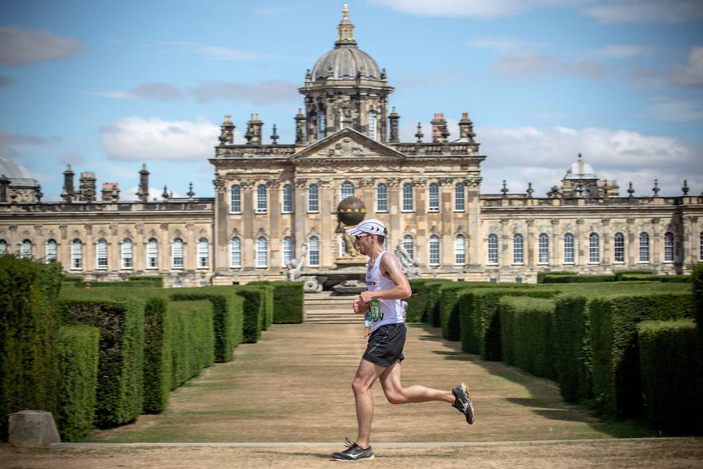 A person dressed in athletic gear, including a white cap and sunglasses, is running along a manicured garden pathway with neatly trimmed hedges on both sides. In the background, a grand and ornate historical building with a large dome is visible under a partly cloudy sky.