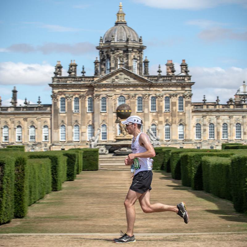 A person dressed in athletic gear, including a white cap and sunglasses, is running along a manicured garden pathway with neatly trimmed hedges on both sides. In the background, a grand and ornate historical building with a large dome is visible under a partly cloudy sky.