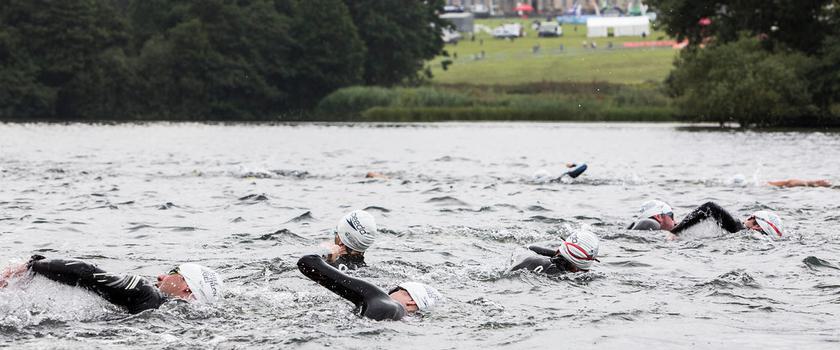 A group of swimmers in wetsuits and swim caps swim in open water during a triathlon event. The background features a large historic building on a hill with lush green trees surrounding the area. The sky is overcast.