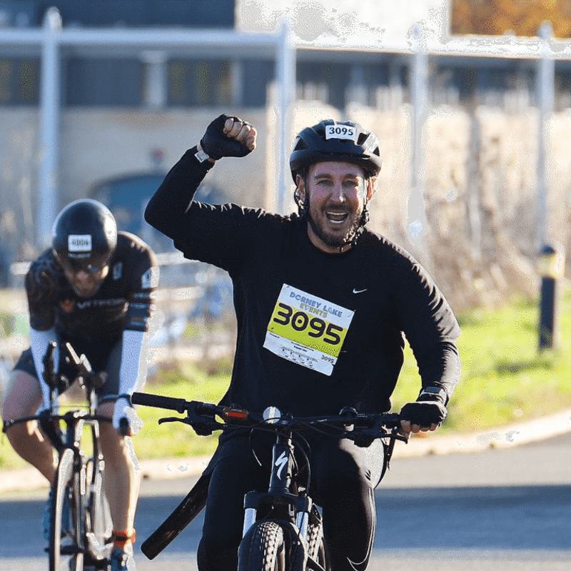 A cyclist in a race, wearing a helmet and a bib numbered 3095, triumphantly raises a fist while riding on a sunny day. Another cyclist follows closely behind, both on a paved path with a building in the background.