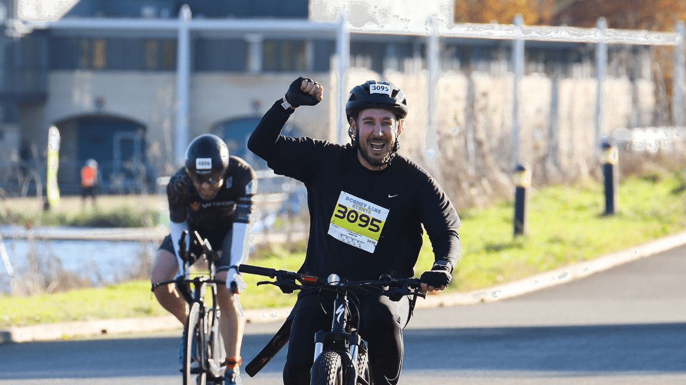 A cyclist in a race, wearing a helmet and a bib numbered 3095, triumphantly raises a fist while riding on a sunny day. Another cyclist follows closely behind, both on a paved path with a building in the background.