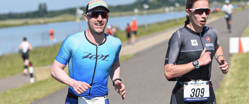 Two athletes are running on a paved pathway in a triathlon. The man on the left wears a blue suit and cap, while the woman on the right is in a black and gray suit with sunglasses. Both have race bibs and are focused, with a body of water in the background.