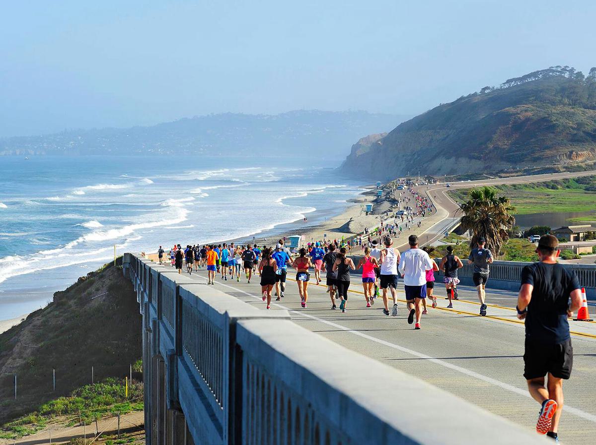 A group of people runs a marathon on a scenic coastal road with views of the ocean and distant hills. The sky is clear, and waves crash on the beach below. Participants wear various athletic outfits as they move along the paved pathway.
