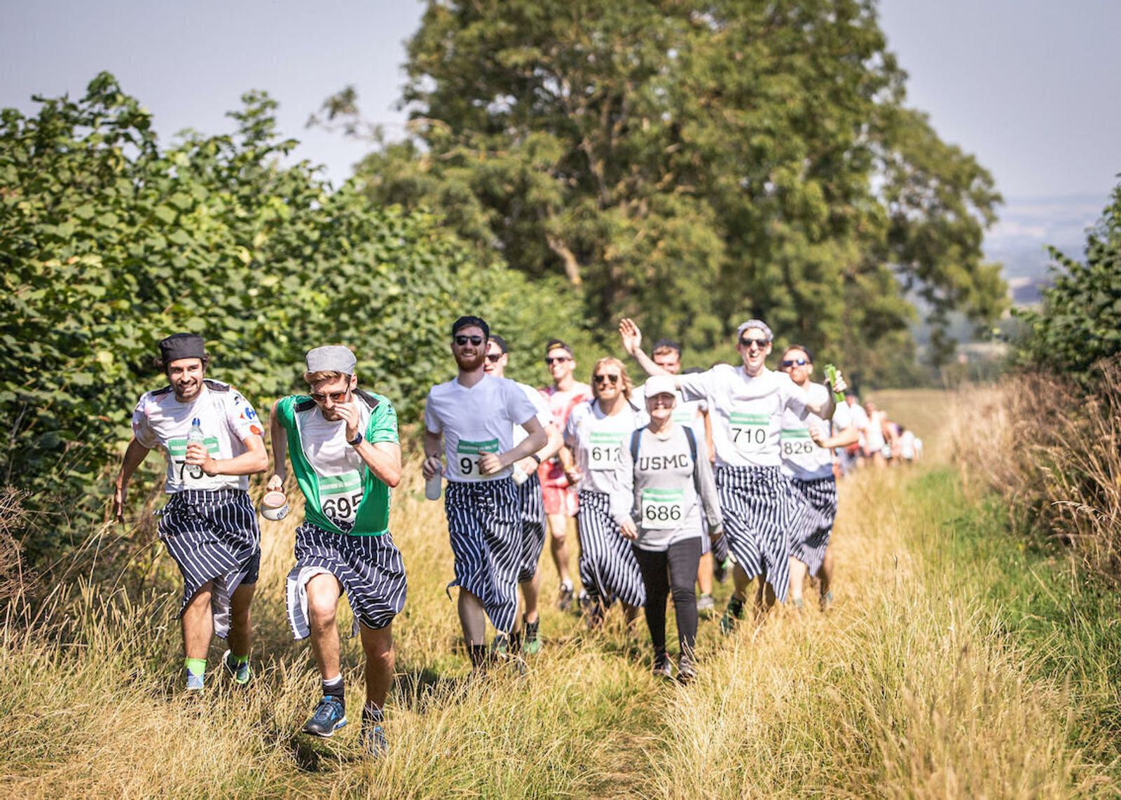 A large group of people, some dressed in similar striped shirts and chef hats, running on a grassy path surrounded by trees and bushes. They all have race bibs pinned to their shirts and appear to be participating in a fun run or charity event.