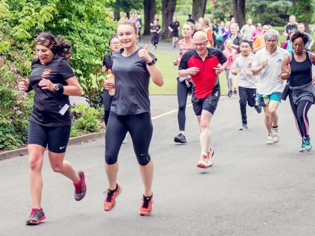 A diverse group of people jogging on a paved path in a park. Participants include both men and women of various ages, wearing athletic clothing. Some runners are smiling or giving thumbs up, with greenery and other joggers visible in the background.