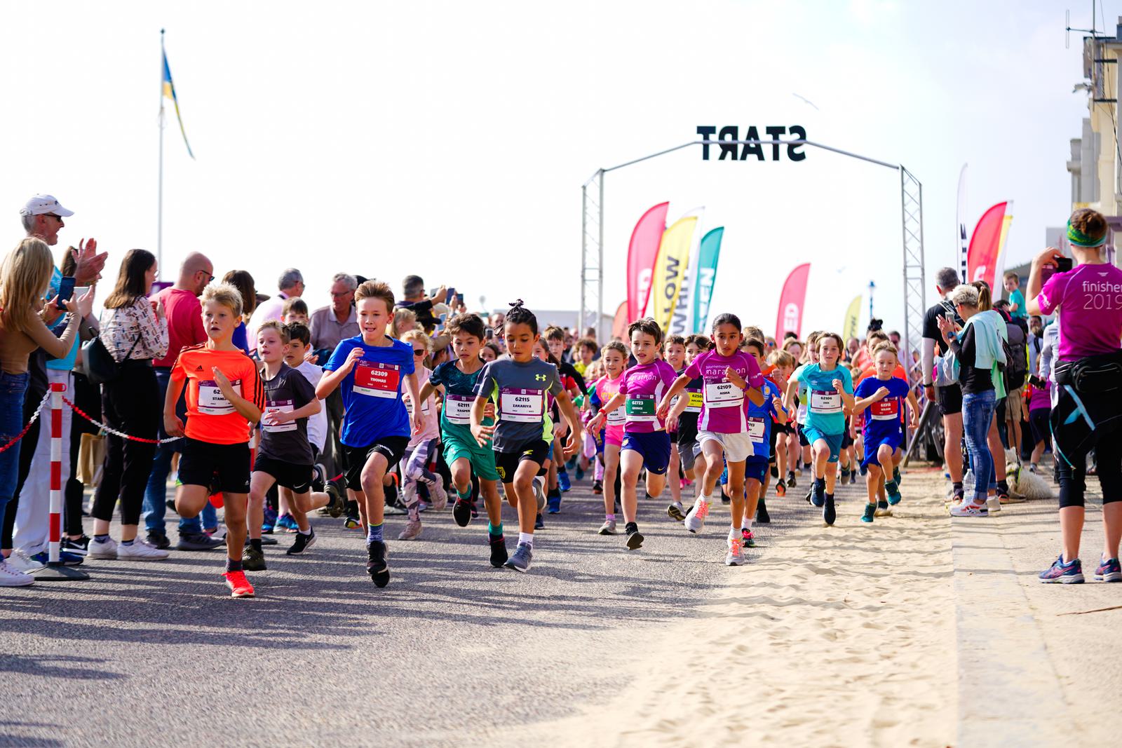 A group of children wearing colorful athletic gear are enthusiastically running at the start of a race. They are on a coastal pathway lined with spectators who are cheering them on. Various flags and race markers are visible in the background.