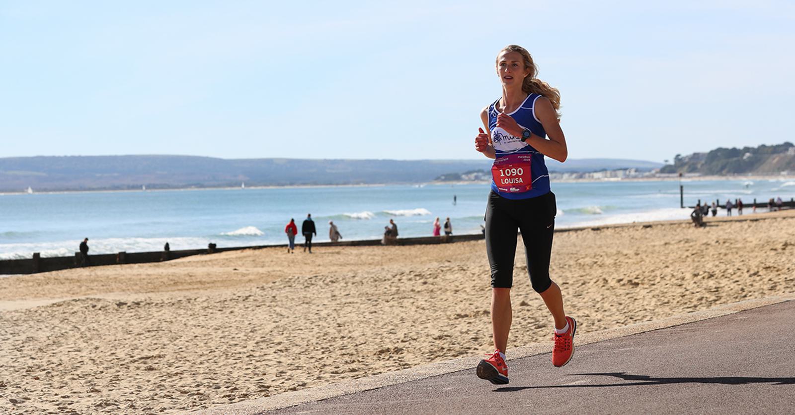 A woman running along a beachfront path wearing a blue tank top, black leggings, and red shoes. She has a race bib with the number 1090. The beach and ocean are in the background, with other people walking in the distance on a sunny day.