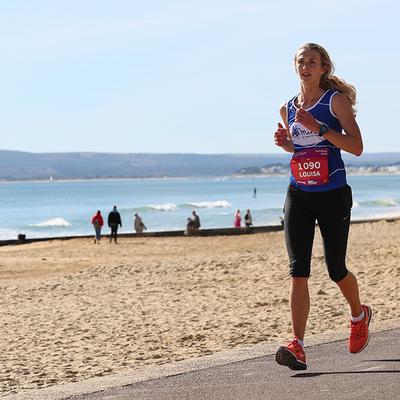 A woman running along a beachfront path wearing a blue tank top, black leggings, and red shoes. She has a race bib with the number 1090. The beach and ocean are in the background, with other people walking in the distance on a sunny day.