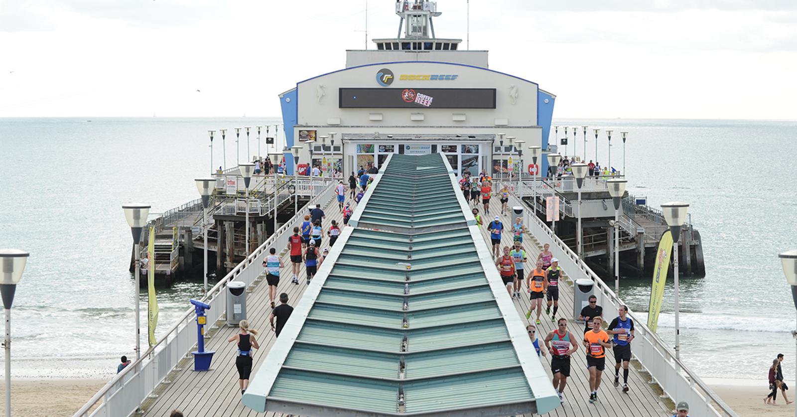 Participants run along a pier towards the ocean, taking part in a race event. The pier stretches out into calm water, and a large building is visible at the end. The sky is overcast, and some spectators are standing along the sides, watching the runners.