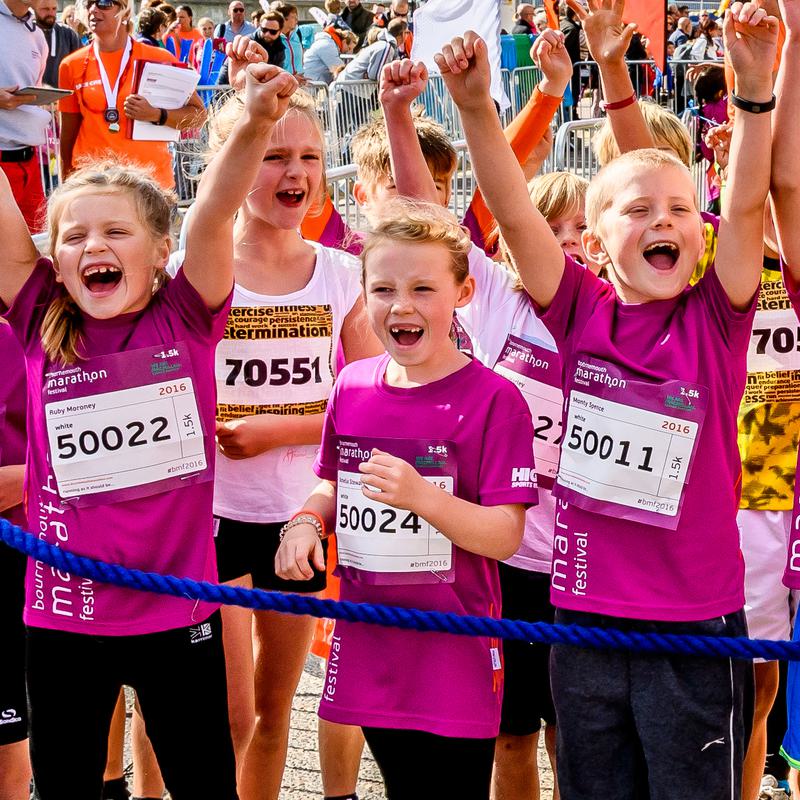 A group of excited children in sportswear cheer with their arms raised at a running event. They wear pink shirts and race bibs and stand behind a blue rope barrier, surrounded by spectators and other participants.