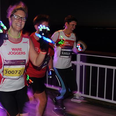 Three runners participate in a night race, identifiable by their illuminated gloves and ware joggers attire. The background features a dark, nighttime sky. The runners seem energetic, and one of them flashes a cheerful smile at the camera.