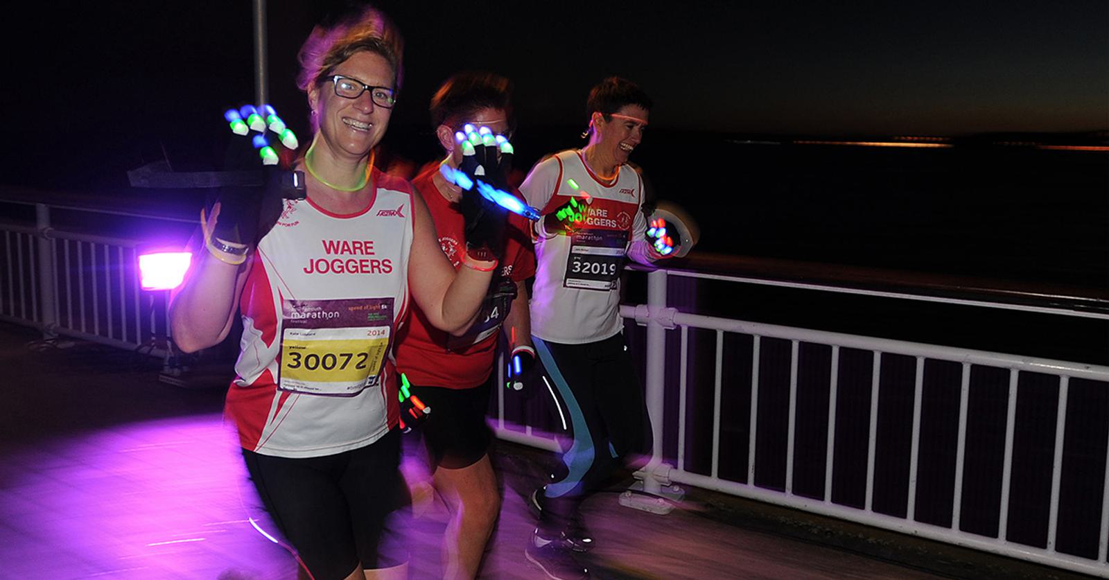 Three runners participate in a night race, identifiable by their illuminated gloves and ware joggers attire. The background features a dark, nighttime sky. The runners seem energetic, and one of them flashes a cheerful smile at the camera.