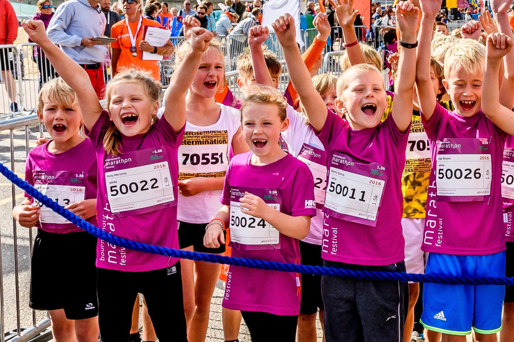A group of excited children in sportswear cheer with their arms raised at a running event. They wear pink shirts and race bibs and stand behind a blue rope barrier, surrounded by spectators and other participants.