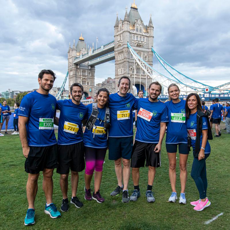 A group of seven people dressed in matching blue athletic wear stands smiling in front of Tower Bridge. They all wear race bibs and pose together on a grassy area, suggesting participation in a running event. Other participants and the Thames River are visible in the background.