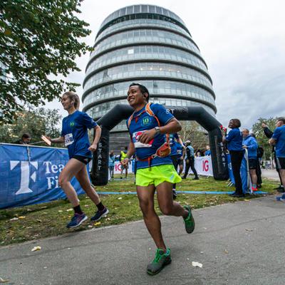 Two runners, wearing colorful athletic gear, participate in a race near a modern, rounded building. A photographer captures the moment nearby. Other participants and onlookers are visible in the background, and trees line the path.