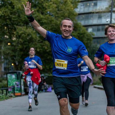 A group of runners is participating in a race. They are wearing blue shirts with numbers, running on a path through a park. A man in front, with a yellow bib number 804, is smiling and waving at the camera. Trees and buildings are visible in the background.