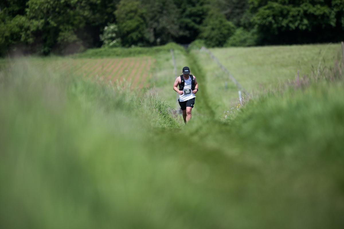 A person in athletic gear and a cap is running on a narrow dirt path through a green, open field, surrounded by lush grass and trees in the background. The focus is on the runner, and the scene is set in a rural area.