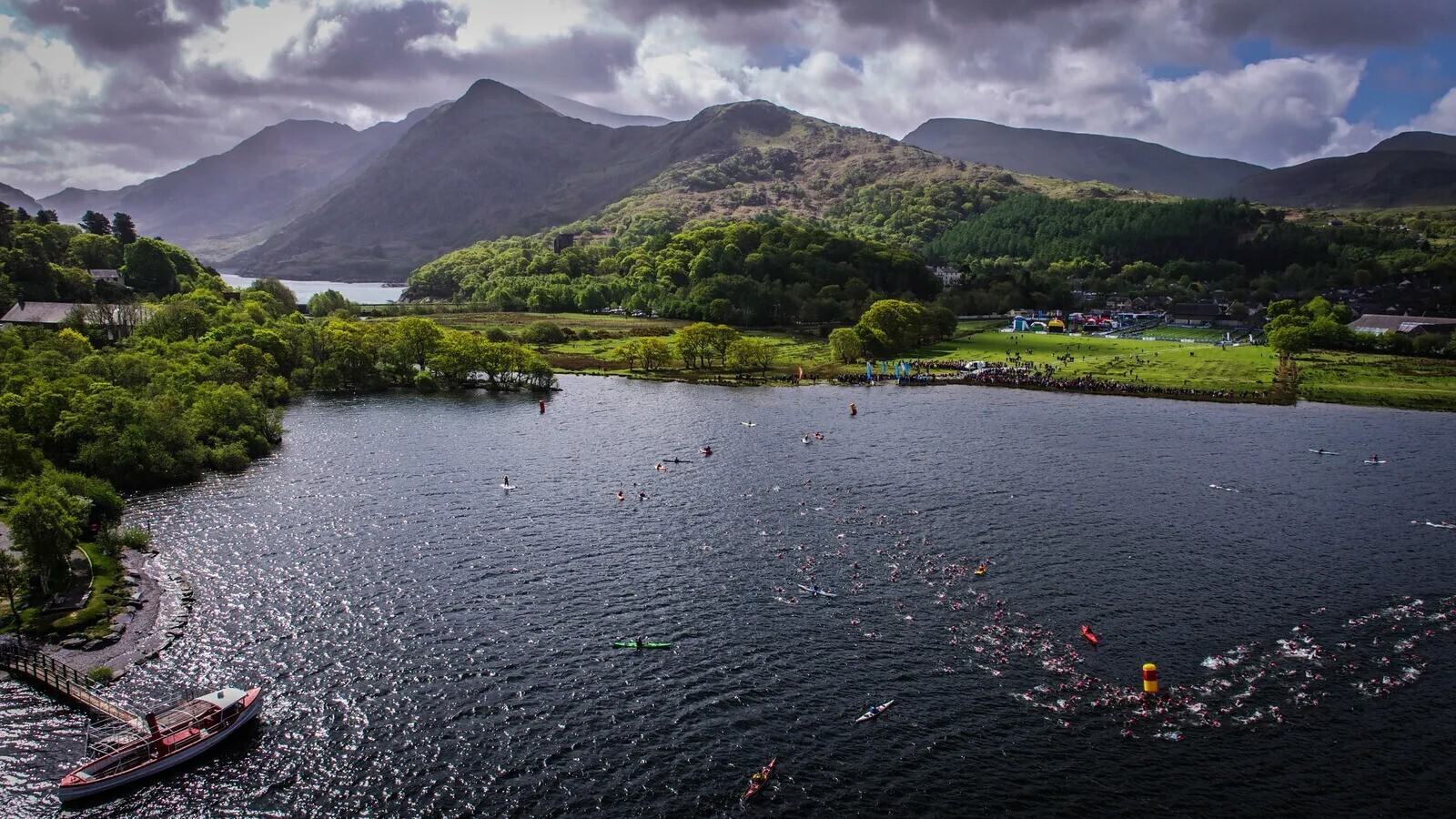 A scenic lake is surrounded by lush green hills and mountains under a cloudy sky. In the water, many swimmers are participating in a race, marked by buoys. On the shore, there is a dock with a boat and a small, vibrant gathering area with tents and colorful flags.