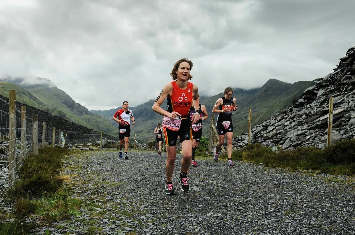 A group of athletes, wearing race bibs and triathlon gear, runs on a gravel trail surrounded by mountainous terrain under a cloudy sky. The lead runner smiles as they advance, with other competitors following closely behind.