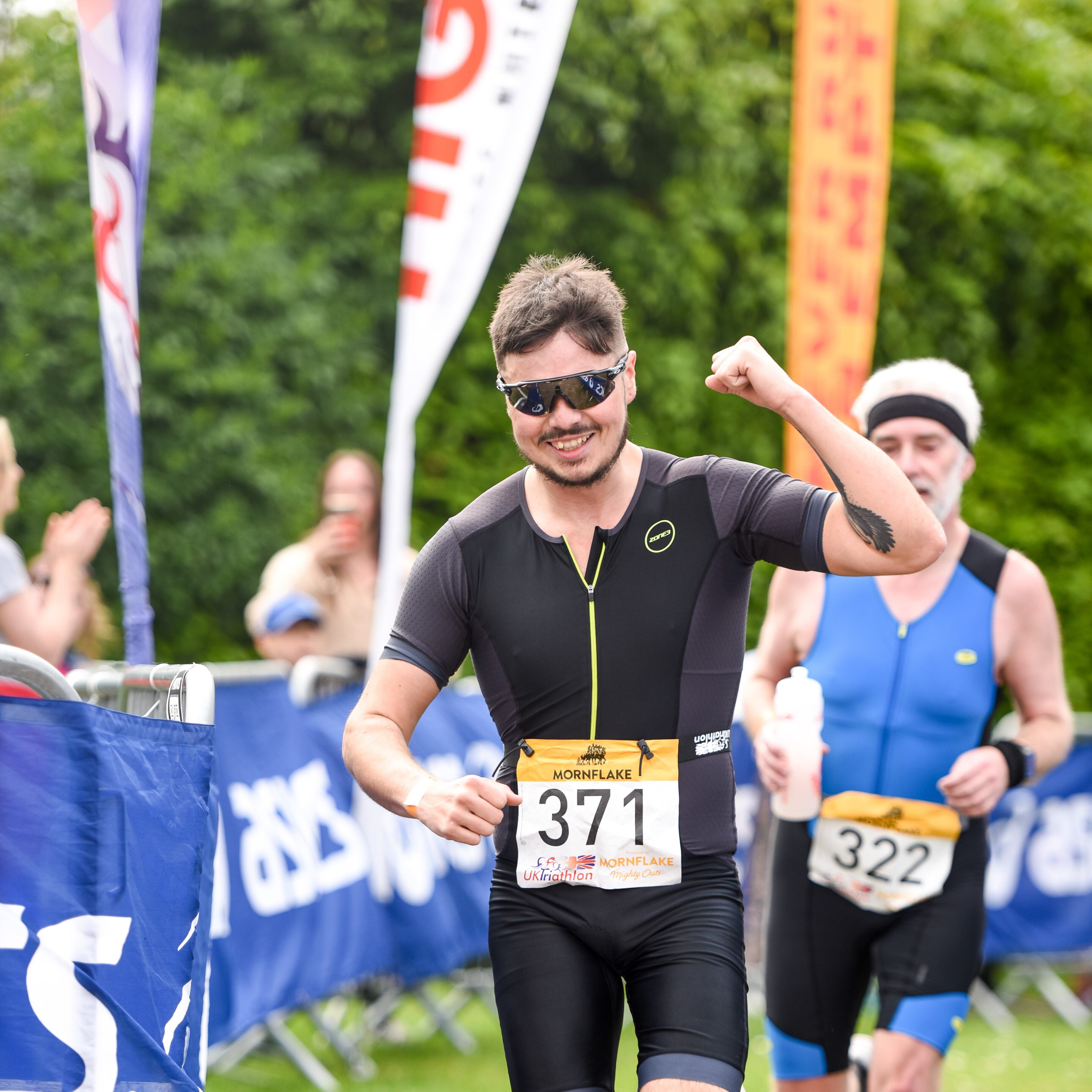 A male athlete wearing sunglasses and a black athletic suit with the number 371 on his chest celebrates by flexing his arm as he nears the finish line of a race. Another participant in a blue suit with the number 322 is seen in the background. Banners and spectators are visible.
