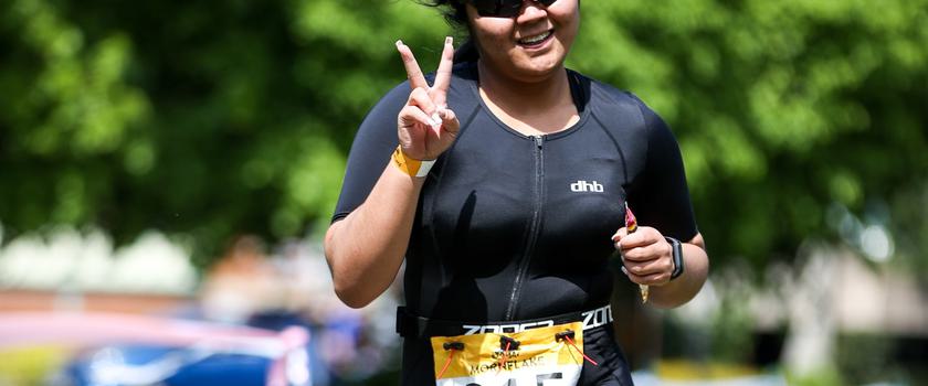 A person wearing sunglasses and a black athletic outfit is running outdoors, smiling, and giving a peace sign with their right hand. They are wearing a race bib with the number 815. The background features lush green trees and a sunny day.