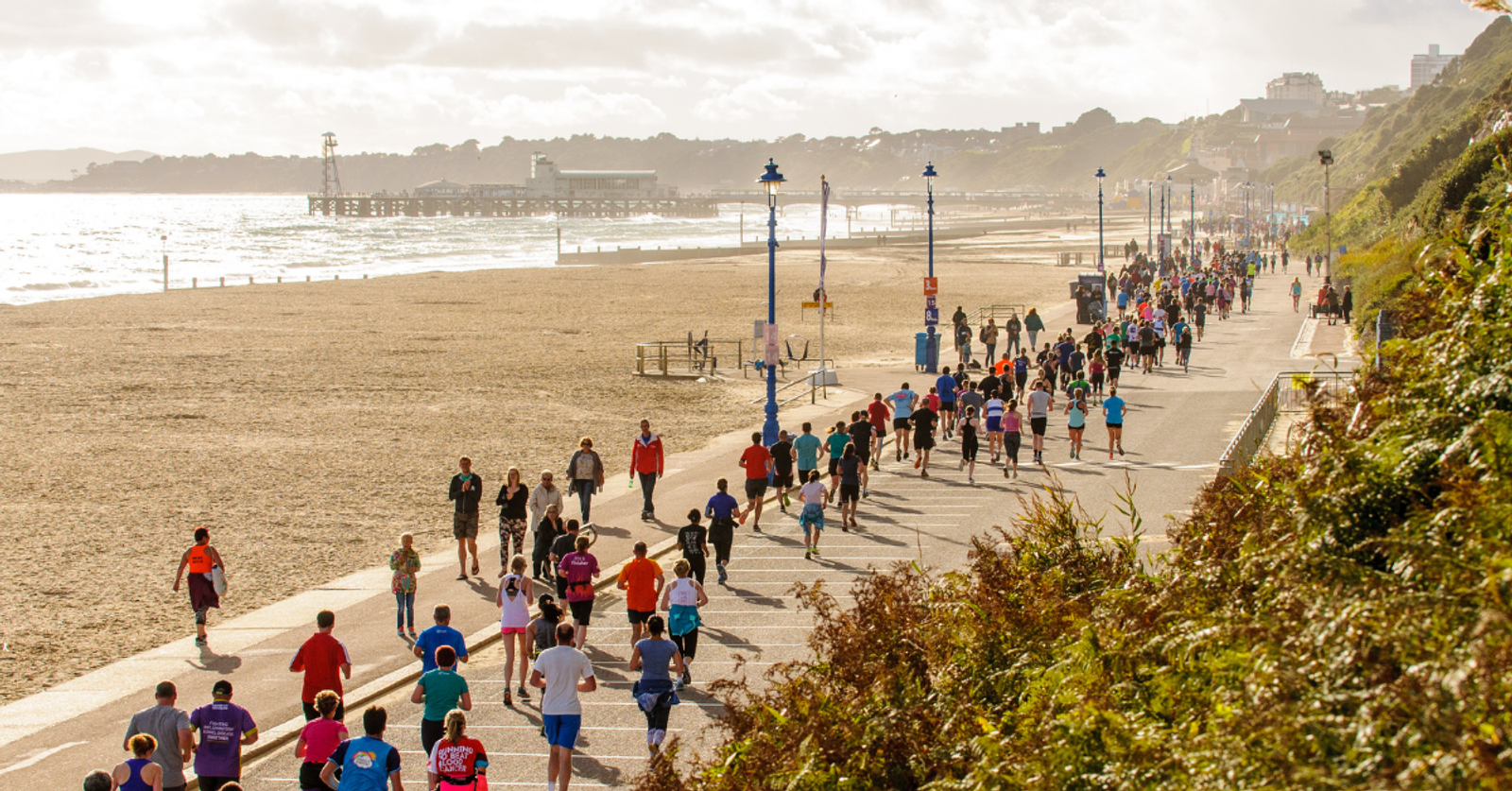 A large group of runners participate in a marathon along a paved seaside promenade, with the beach and ocean to their left. The sky is partly cloudy, and there are buildings and a pier visible in the distance. Green shrubbery borders the path to the right.