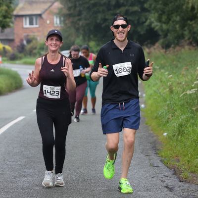Two runners in athletic gear smile and gesture with their hands while jogging on a rural road, surrounded by greenery and other participants in the background. Both are wearing numbered bibs, indicating they are in a race.