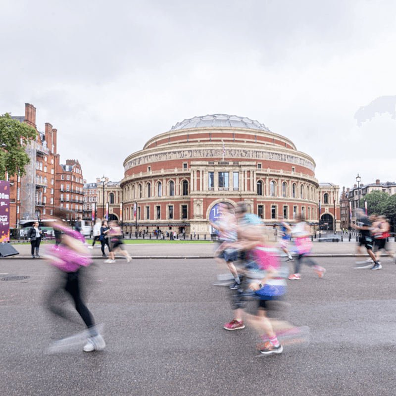 People are running in front of a large, historic building with a domed roof and detailed facade, on a cloudy day. Trees flank the building, and the scene captures motion blur from the runners, suggesting a race or marathon event in progress.