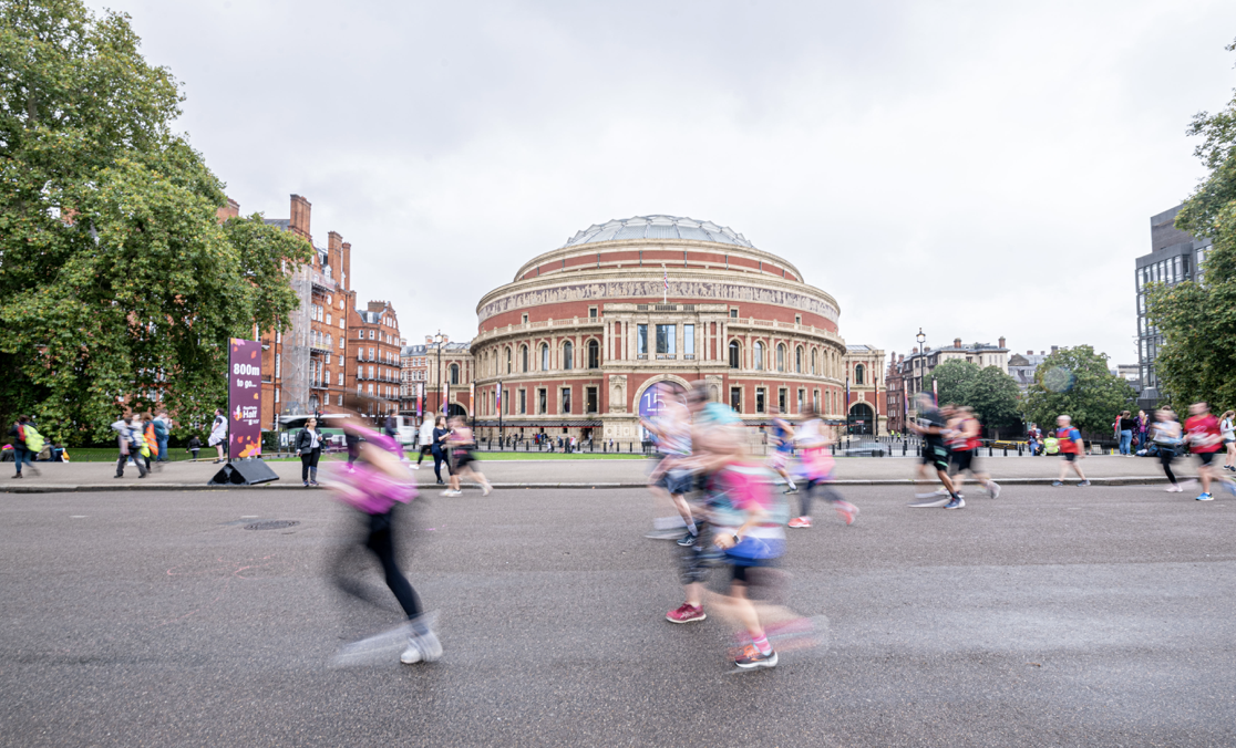 People are running in front of a large, historic building with a domed roof and detailed facade, on a cloudy day. Trees flank the building, and the scene captures motion blur from the runners, suggesting a race or marathon event in progress.