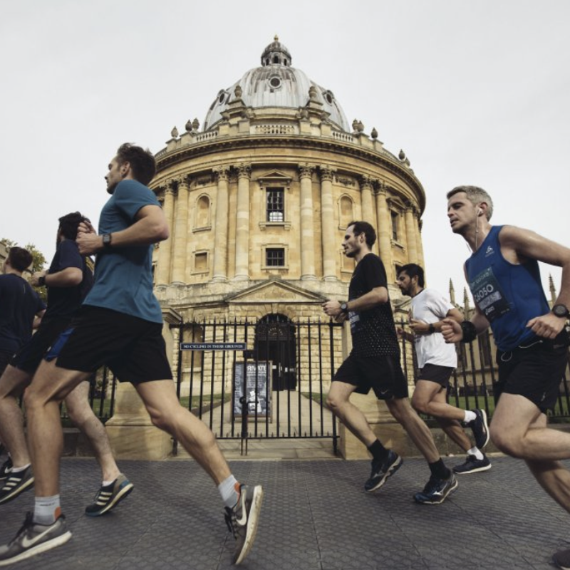 A group of runners jogs in front of the Radcliffe Camera, a historic building in Oxford, England. The runners wear athletic attire and appear to be participating in a race or organized run. The sky is overcast, and the architectural details of the building are prominent.